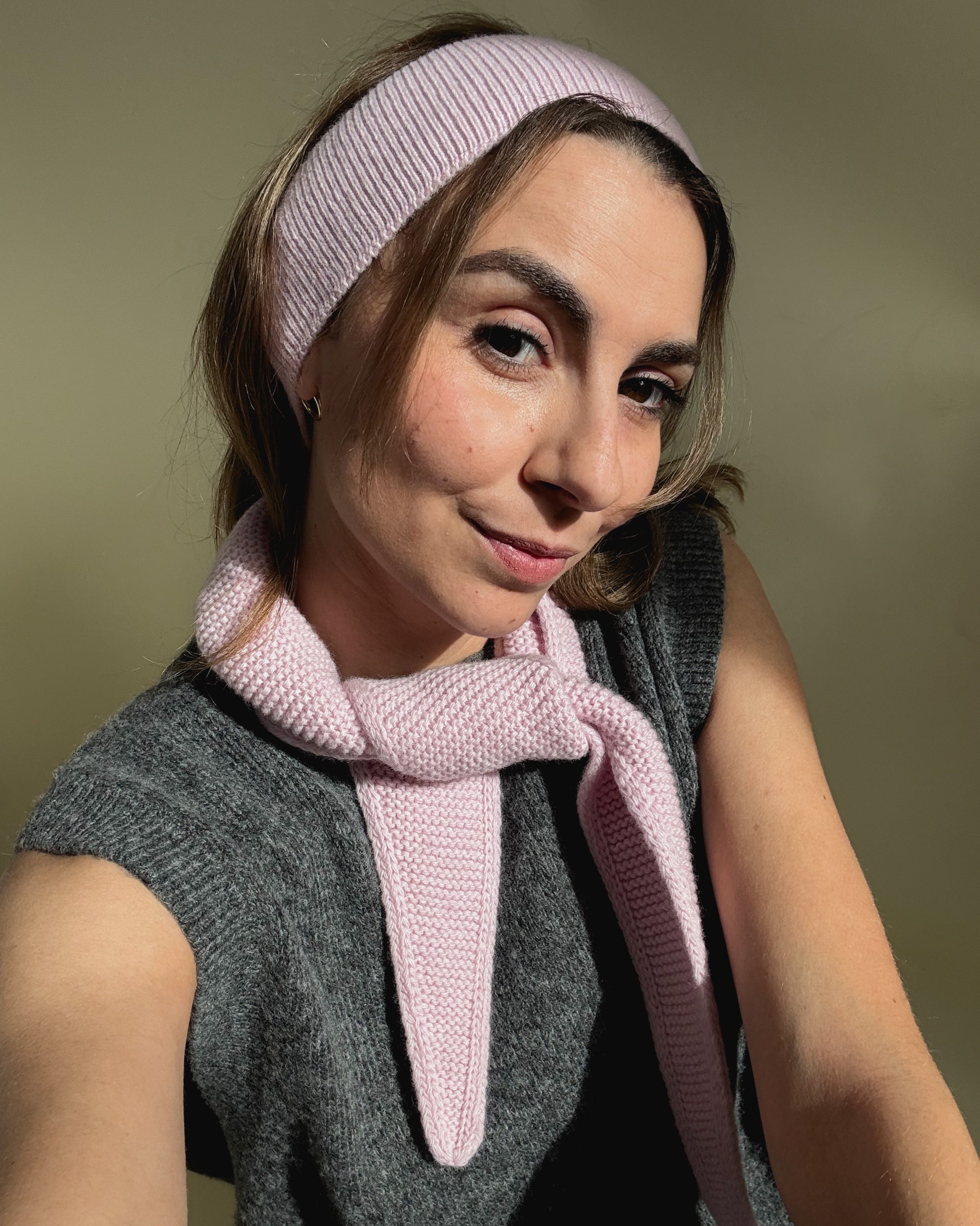 eine frau schaut in die kamera und trägt ein stirnband sowie einen mini scarf aus merinowolle in der farbe pink